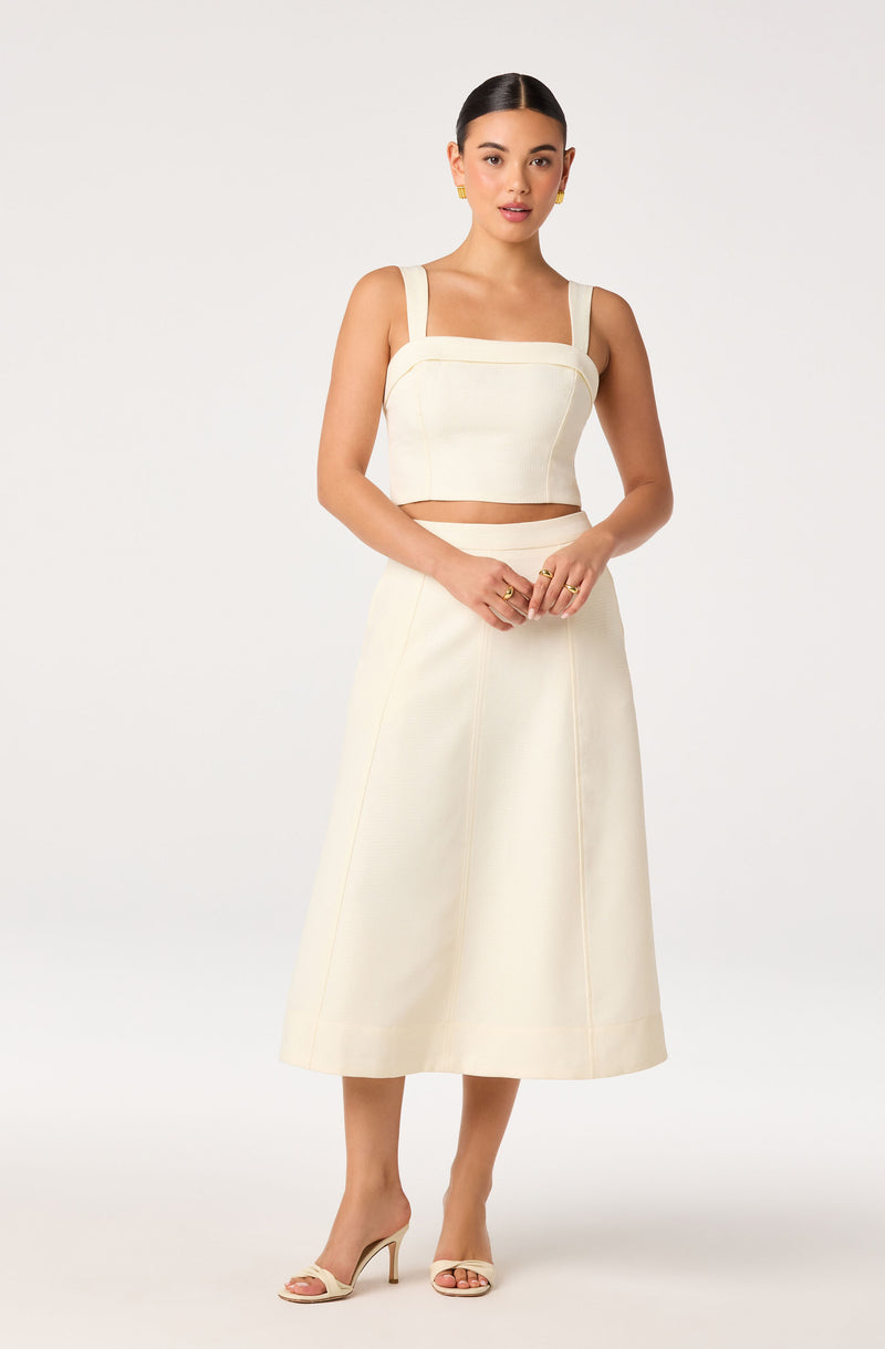 Woman in cream two-piece outfit with wide strap top and A-line skirt, wearing beige heels.