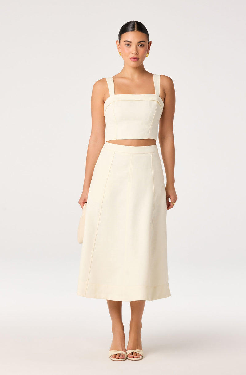 Woman in a cream-colored sleeveless crop top and wide-leg midi skirt, holding a small bag.