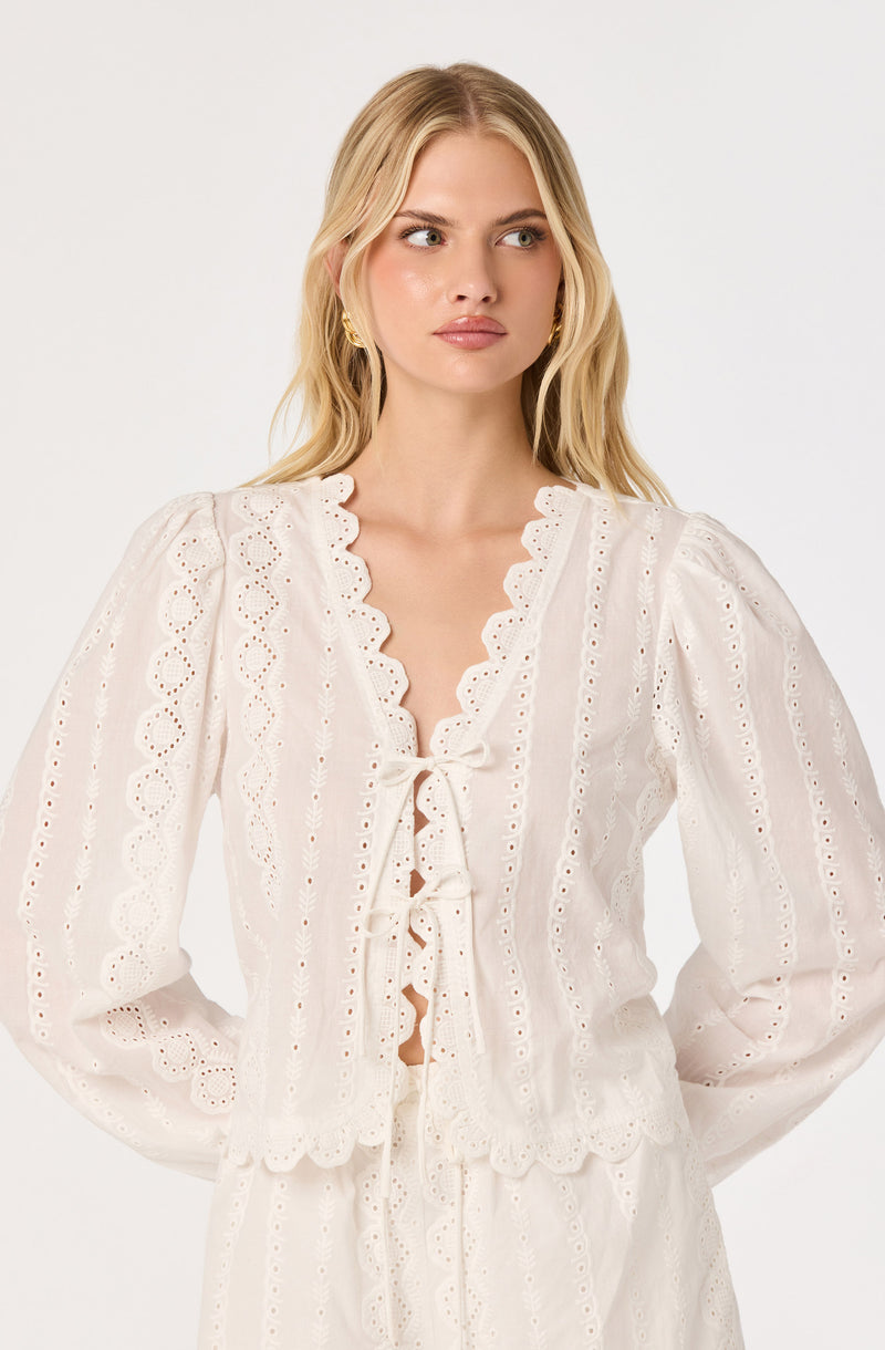 Woman wearing a white eyelet embroidered blouse with ruffled edges and a front tie.