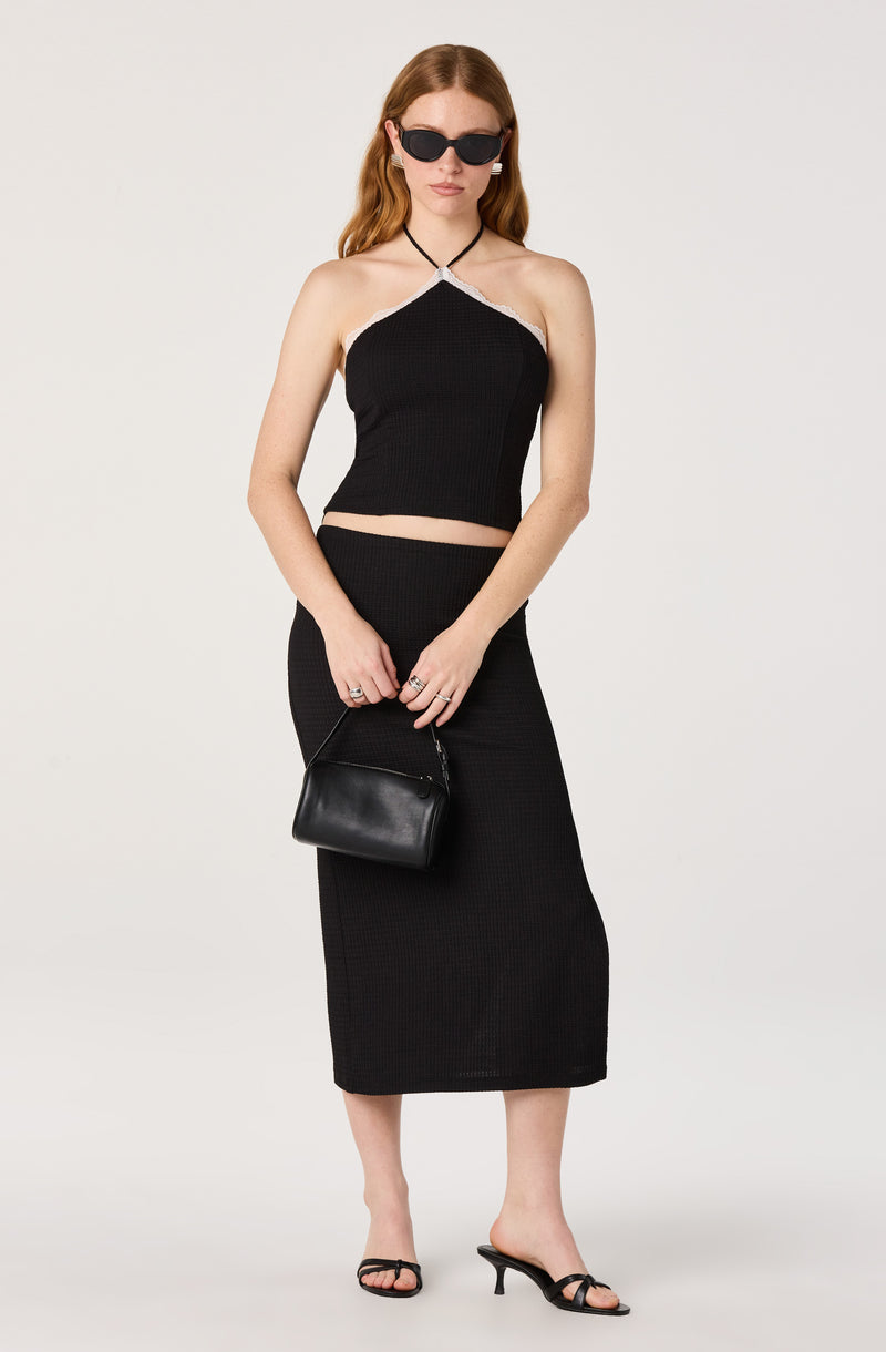 Woman in a black sleeveless halter crop top and matching long skirt, paired with black heeled sandals, sunglasses, and a black handbag.
