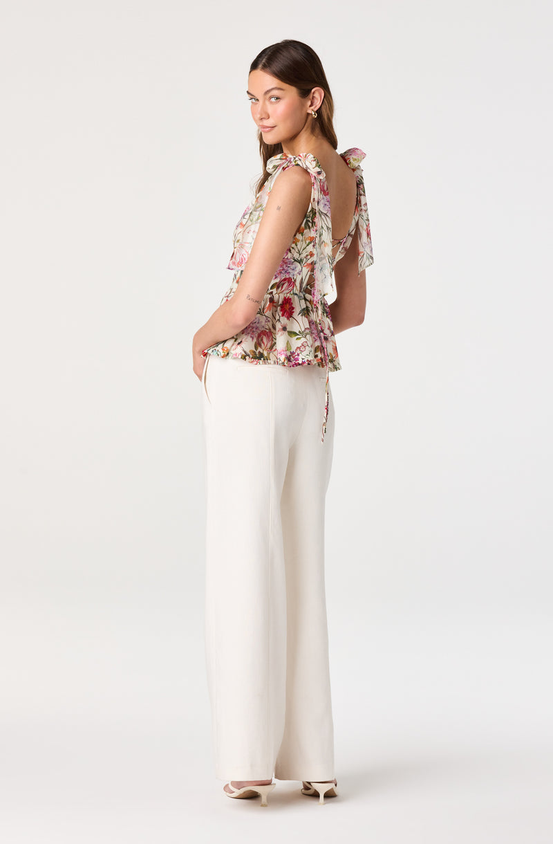 Woman in a sleeveless floral top with ruffled sleeves and bow ties on shoulders, paired with white wide-leg pants.