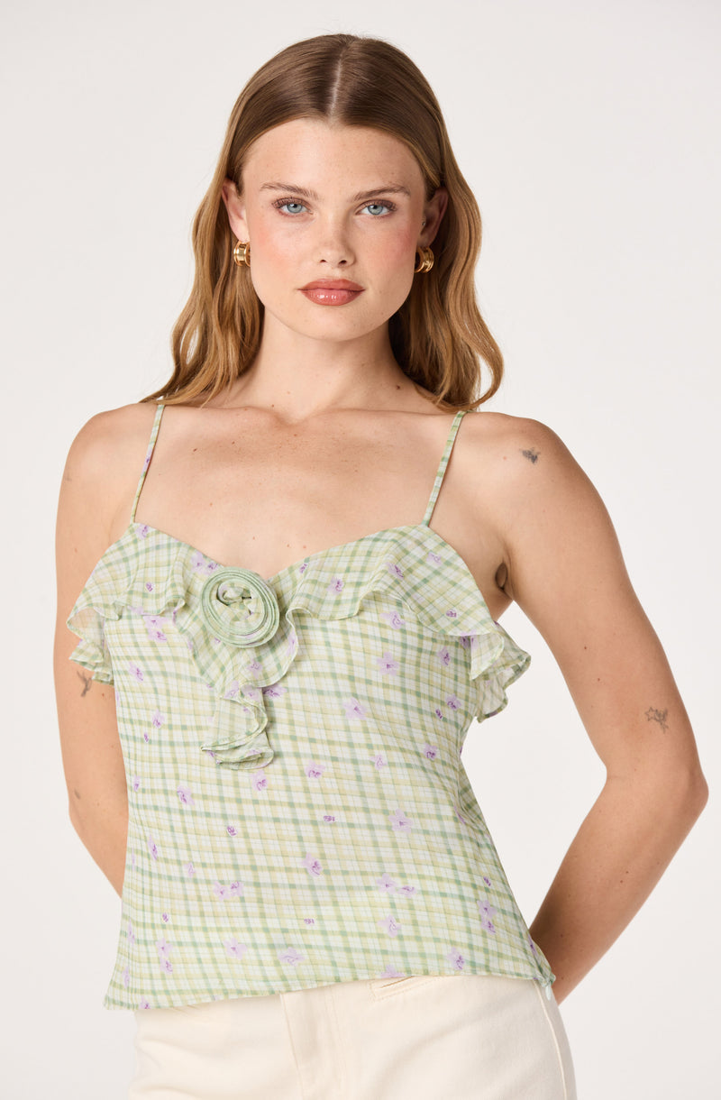 Woman wearing a green and purple checkered sleeveless top with ruffles and a floral brooch detail.