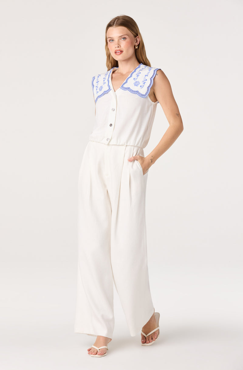 White sleeveless top with blue embroidered collar and buttons, paired with high-waisted white pants.