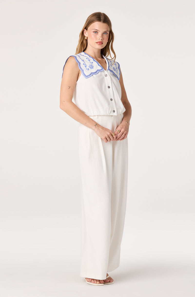 Woman wearing a sleeveless white top with a large blue embroidered collar and wide-leg white pants.