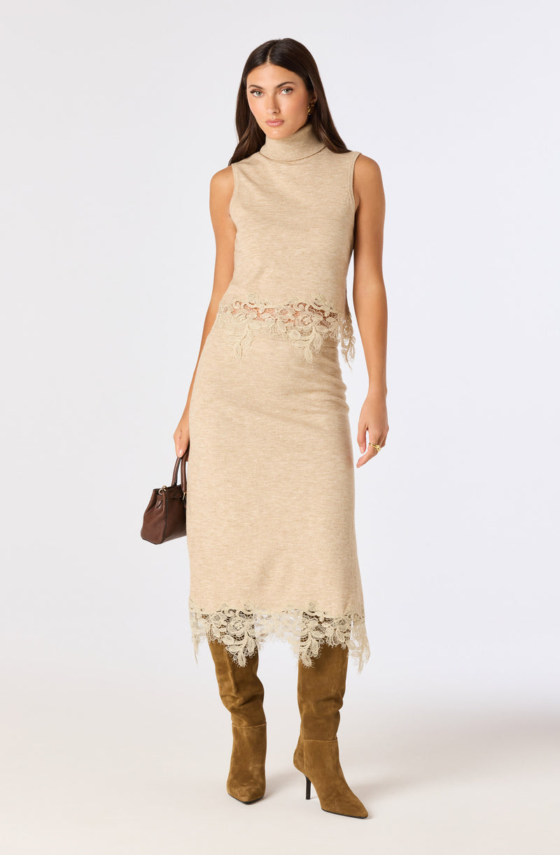 Model wearing a beige sleeveless turtleneck top and a matching beige lace-edged midi skirt, paired with tan knee-high boots.