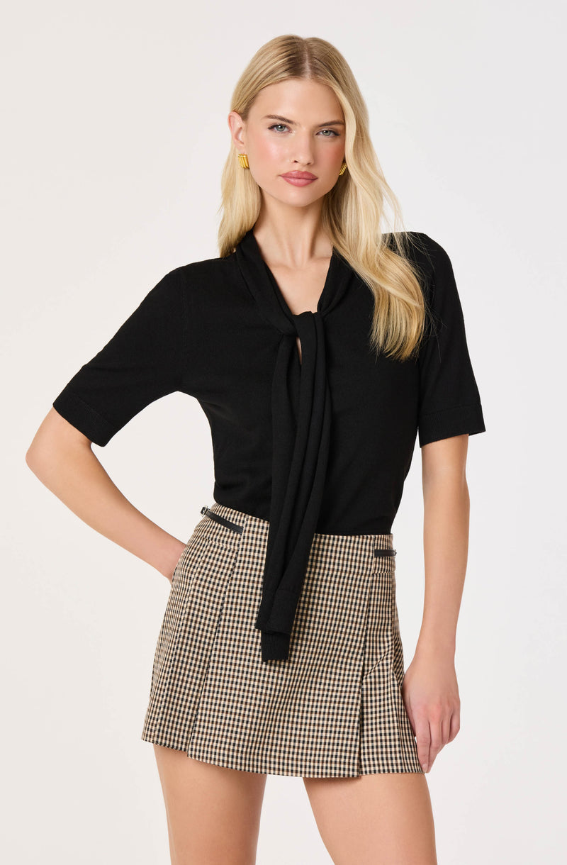 Woman wearing a black top with a neck tie and a beige checkered skirt.
