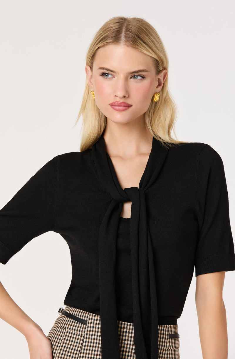 Woman in black top with a necktie, plaid pants, and gold hoop earrings.
