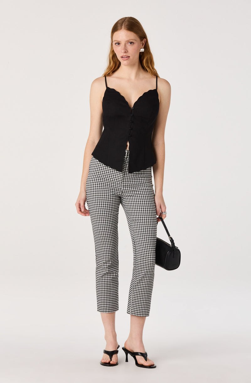 Woman wearing a black sleeveless top with scalloped edges and thin straps, paired with black and white gingham pants and black heels.