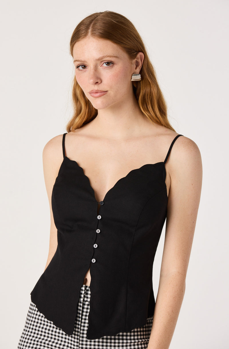 Woman wears a black sleeveless top with scalloped edges and thin spaghetti straps.