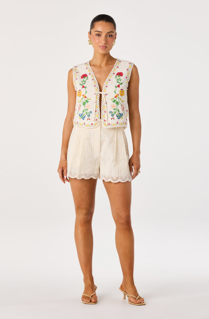 Woman wearing a sleeveless embroidered floral vest and matching eyelet shorts, beige sandals.