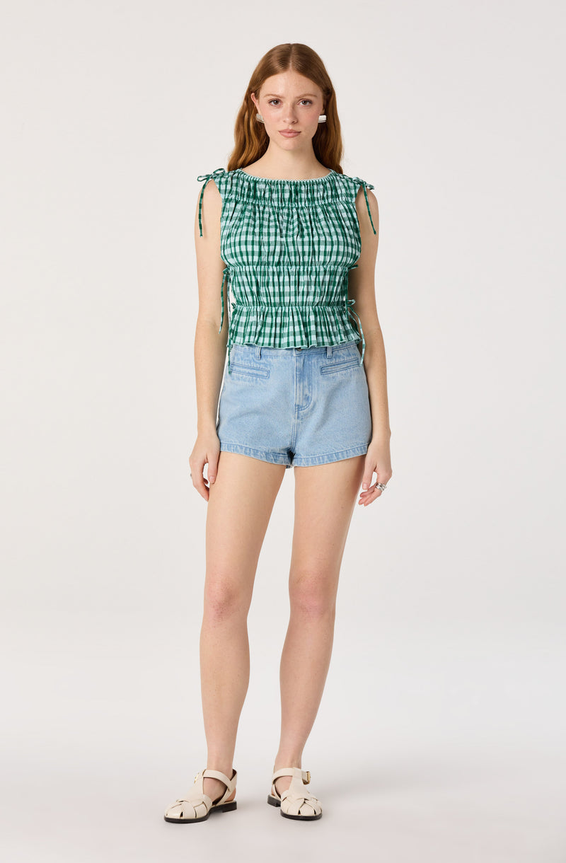 Woman wearing a green and white checkered sleeveless top with ties on shoulders, and light blue denim shorts.