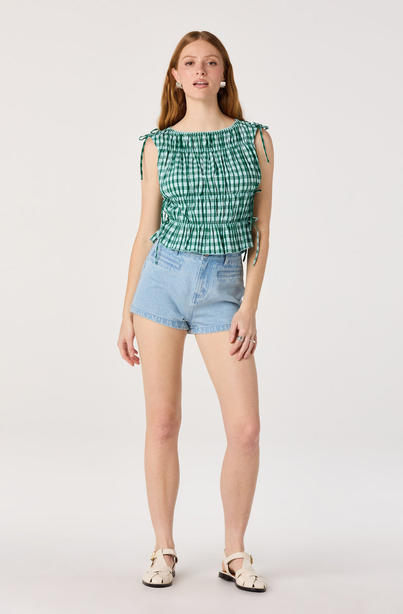 Woman wearing a green and white checkered sleeveless top with drawstring details and light blue denim shorts.