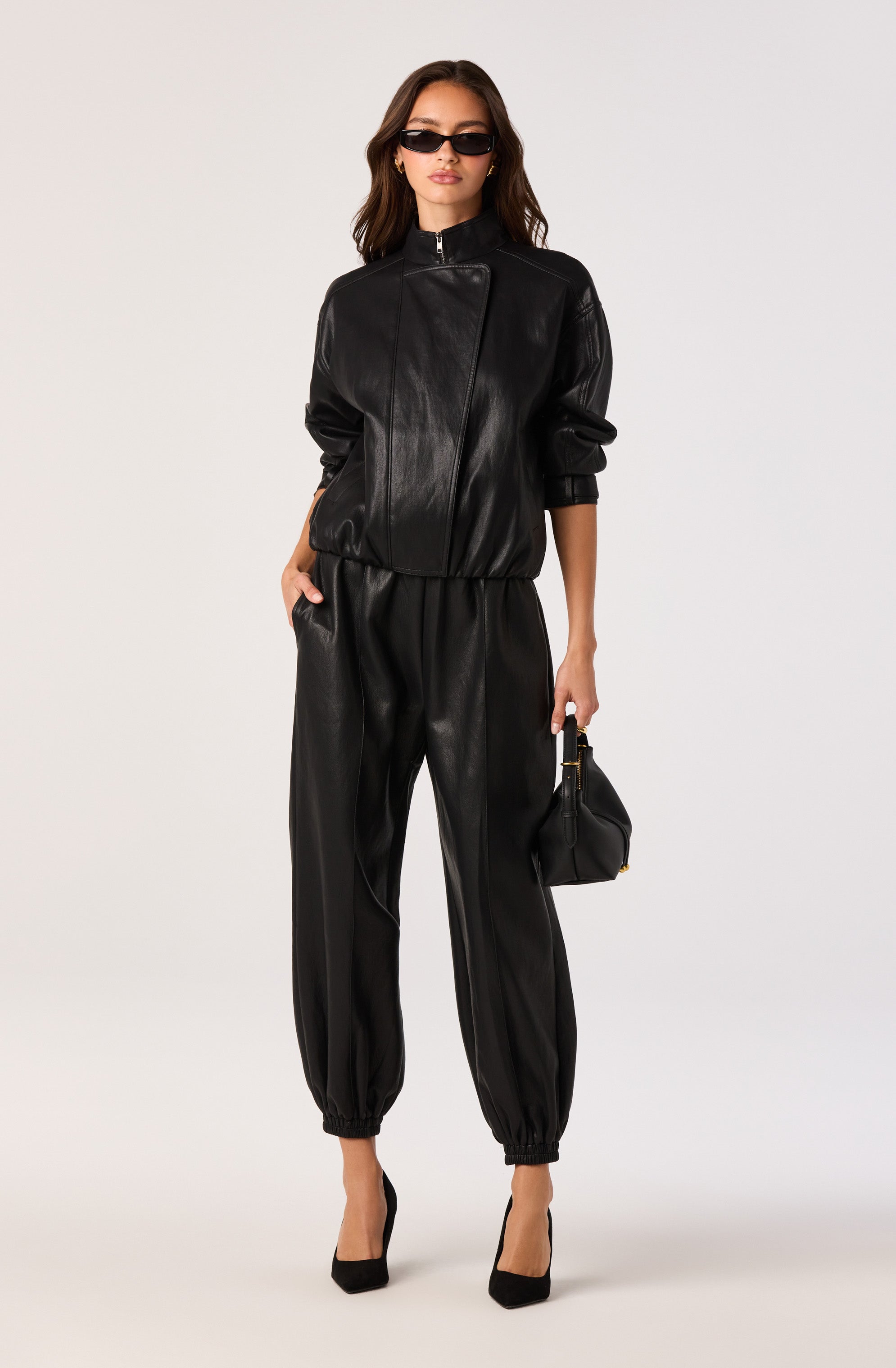 Woman in black leather tracksuit with zip-up jacket, elastic cuffs, and high heels, holding a small black handbag.