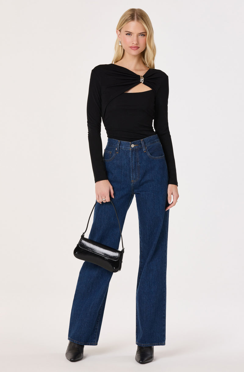 Woman wearing a black twisted-front long-sleeve top with a cutout, high-waisted blue jeans, and black boots, holding a small black handbag.