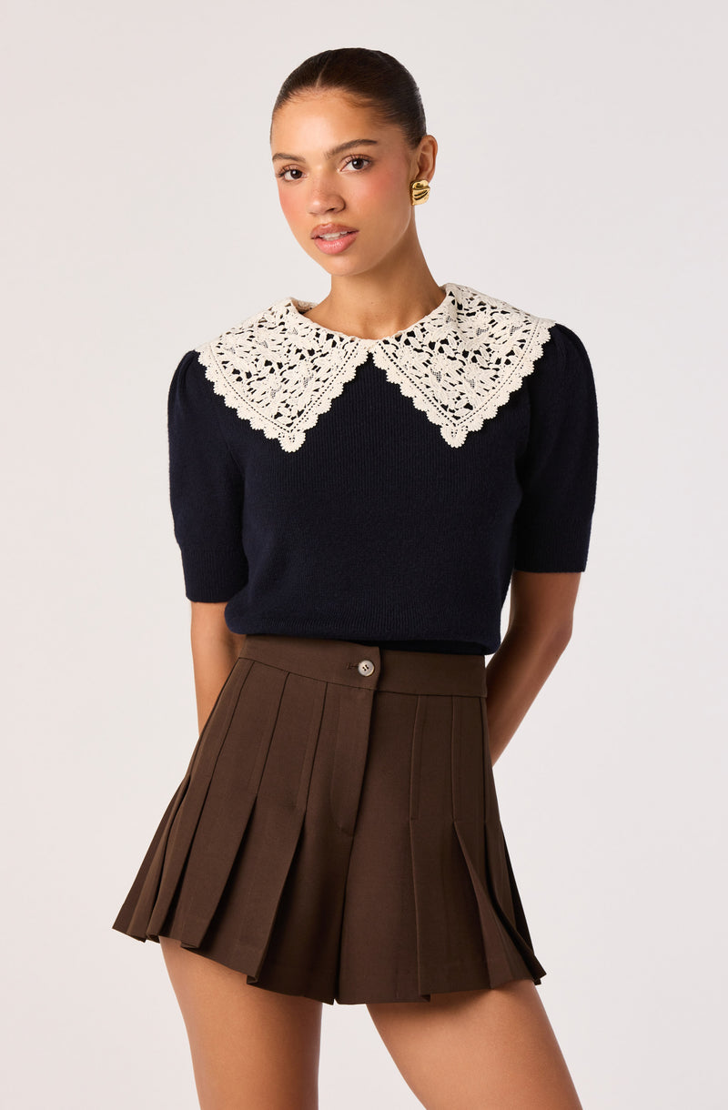 Woman wearing a navy sweater with a large white embroidered collar and brown pleated shorts.
