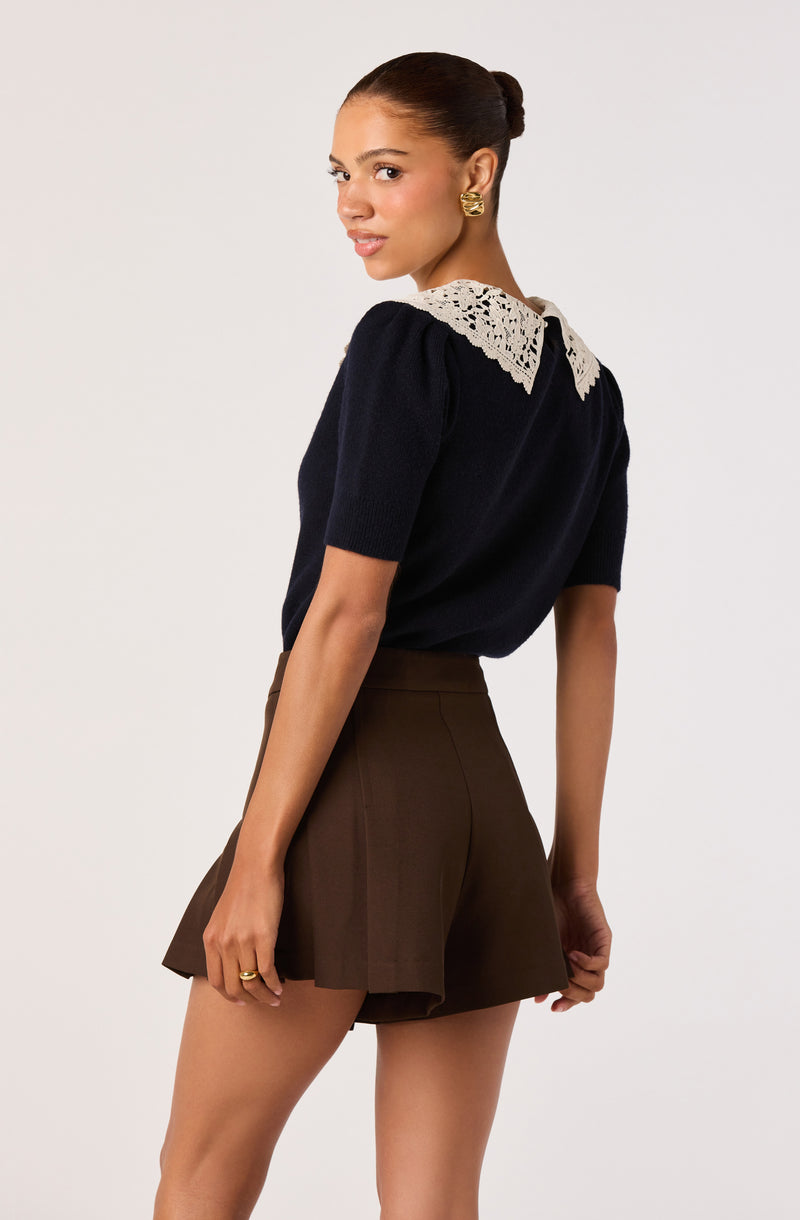 Woman wearing a navy top with a white lace collar and brown pleated shorts.