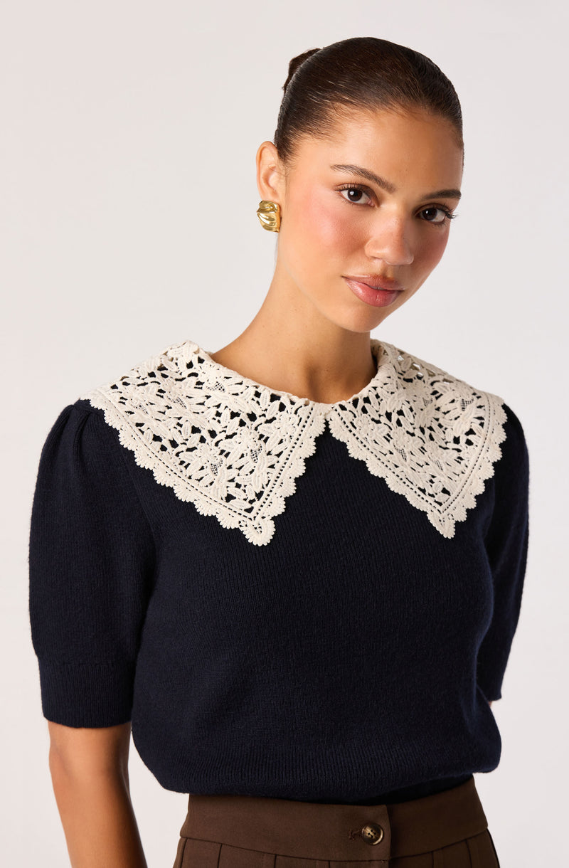 Woman wearing a black sweater with a large cream lace collar and gold earrings.