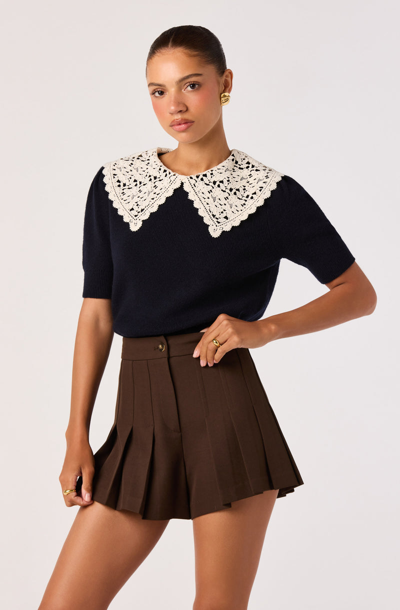 Woman wearing a navy sweater with a large white lace collar and high-waisted brown pleated shorts.