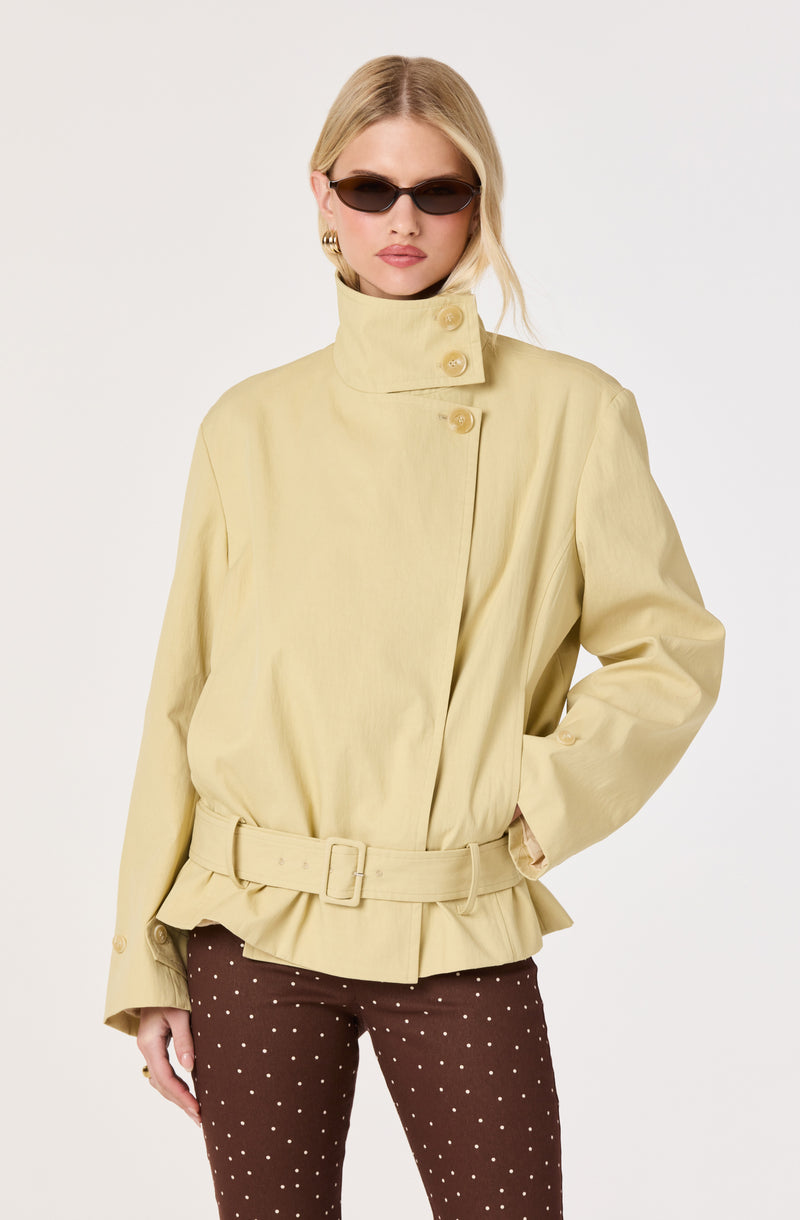 Woman in a beige high-collar jacket with large buttons and a matching belt, paired with brown pants with white polka dots.