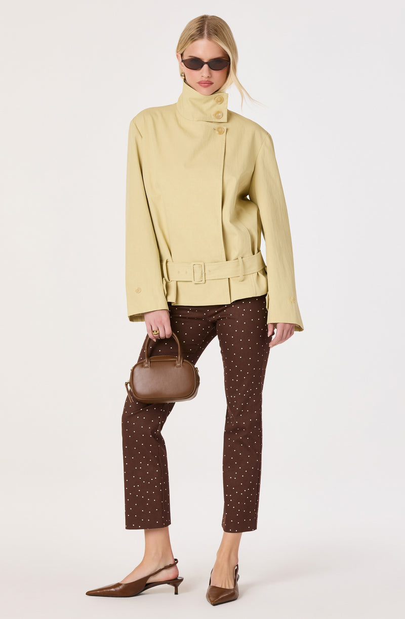 Woman in a yellow high-neck jacket, brown polka dot pants, brown pointed heels, and dark sunglasses, holding a brown handbag.