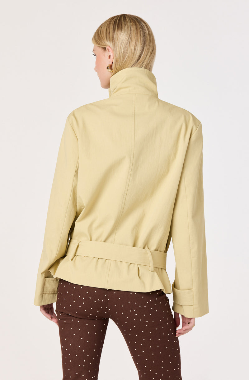 Woman in a beige belted jacket with a high collar and wide polished sleeves, paired with brown pants with white polka dots.