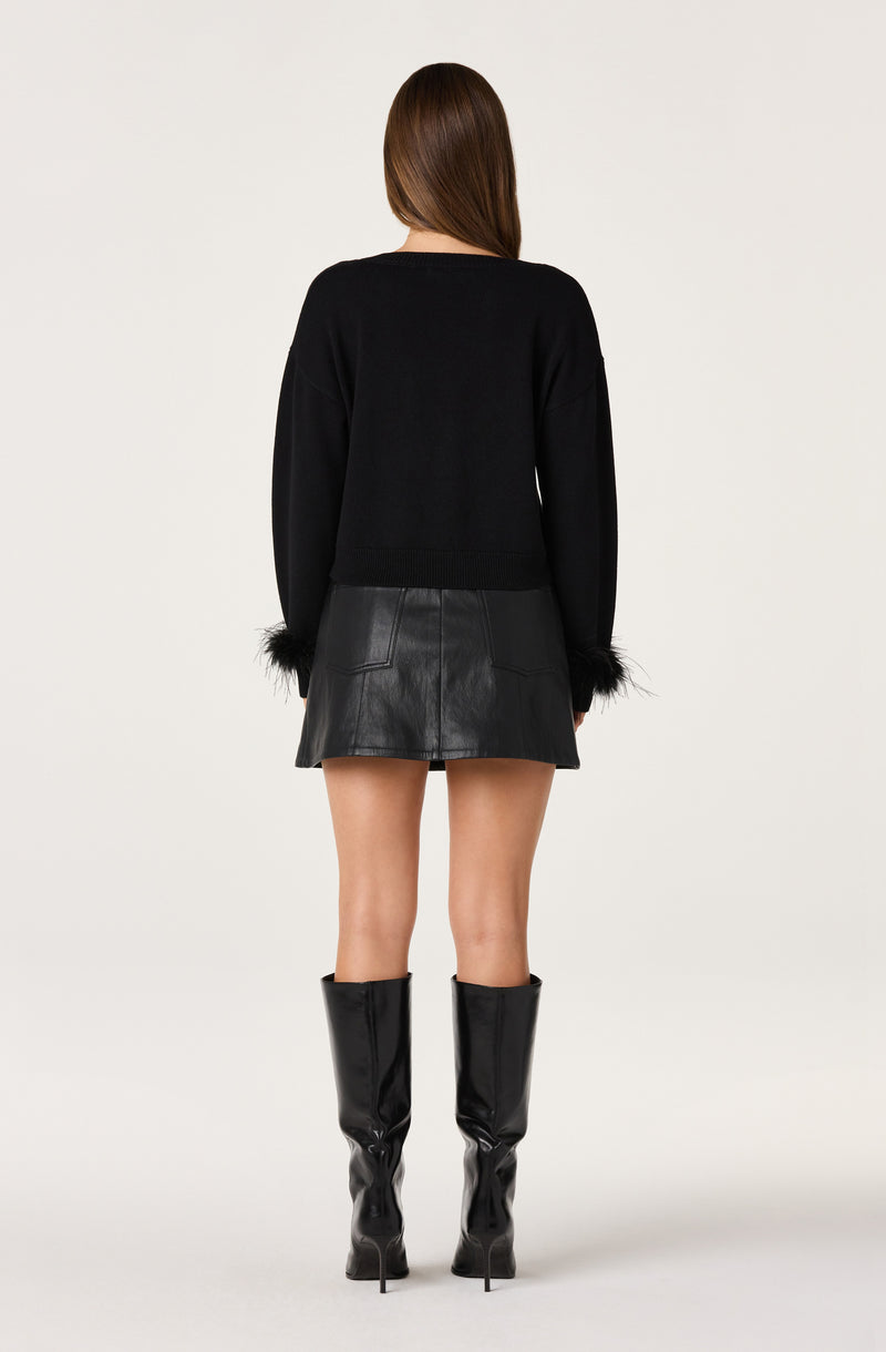 Woman wearing a black long-sleeve sweater with feathered cuffs, a black leather mini skirt, and tall black boots.