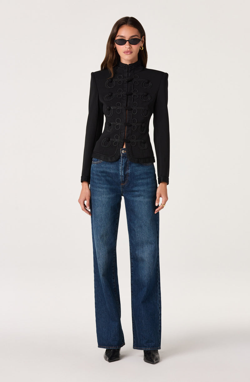 Woman wearing a fitted black jacket with decorative loops, straight-leg blue jeans, black shoes, and sunglasses.