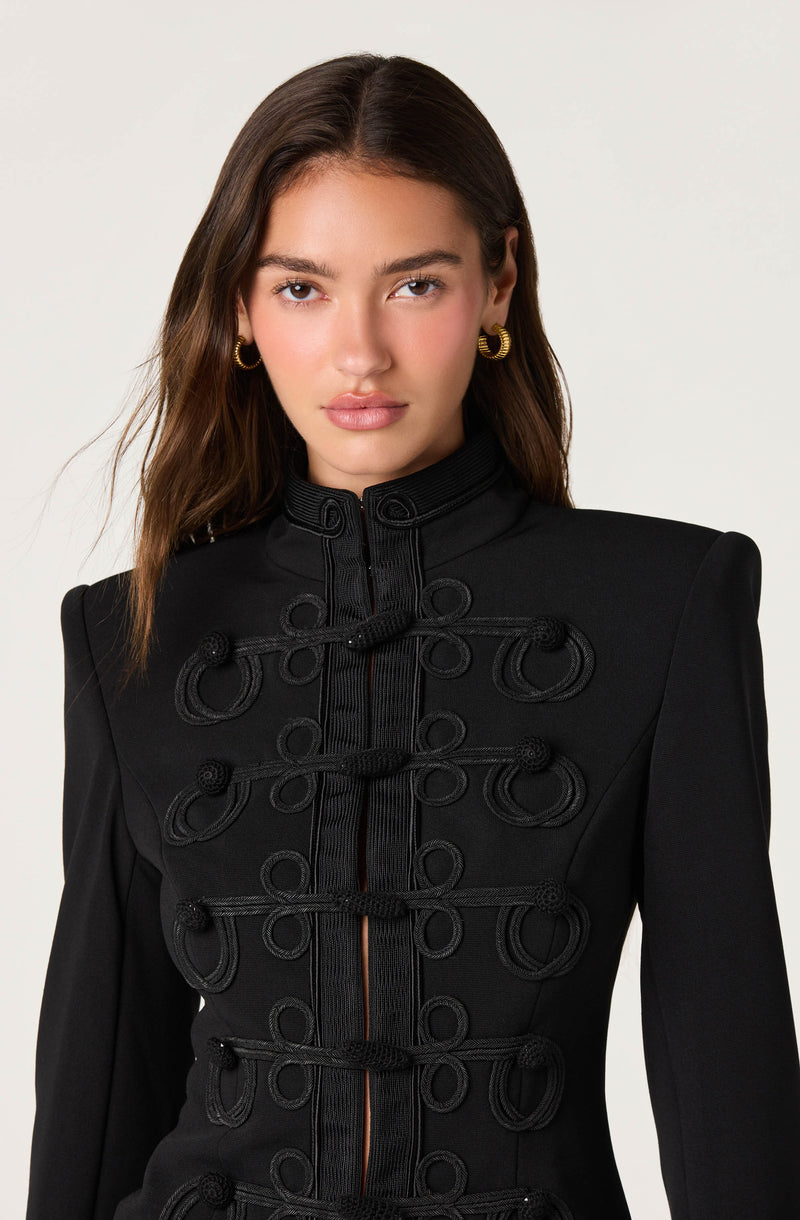 Woman wearing a black military-style jacket with decorative looped cords and a high collar.