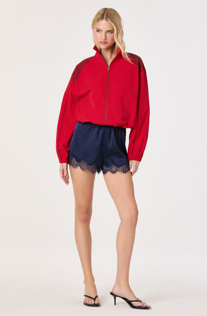 Woman in red and maroon zip-up jacket with navy satin shorts with lace trim and black high heels.