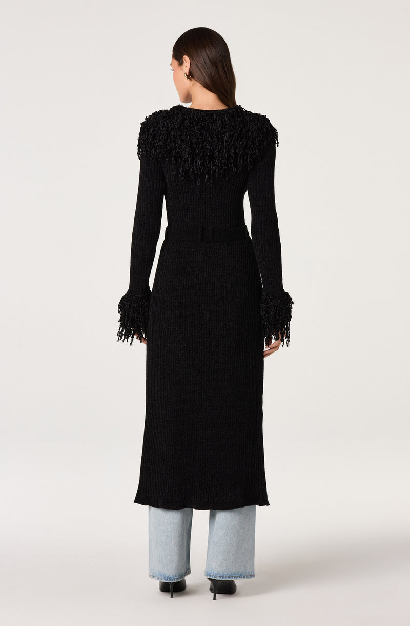 Woman in a long black knit dress with textured shoulders and fringed cuffs, paired with light blue jeans.