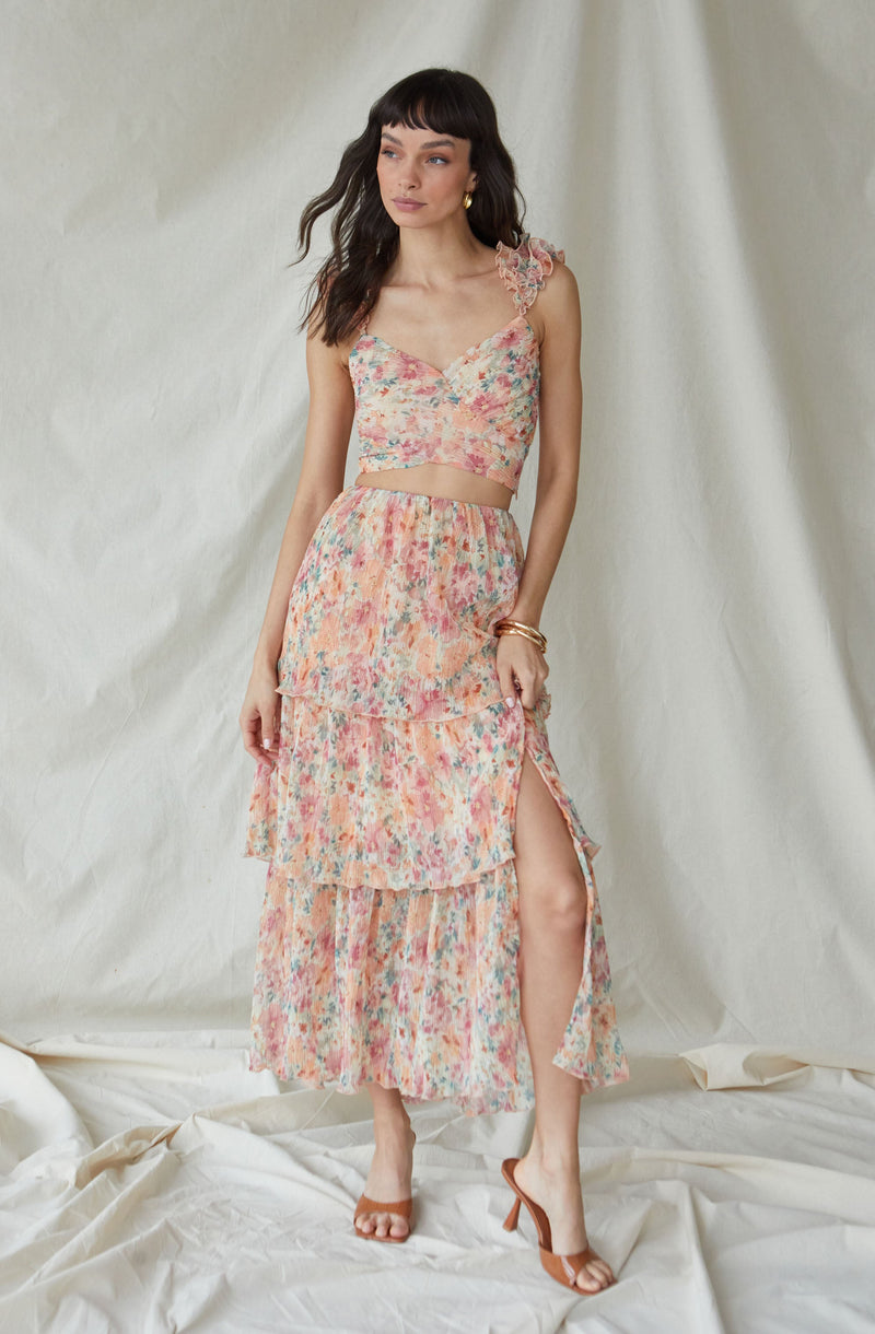 The outfit features a sleeveless crop top with a floral pattern and ruffled shoulder straps. The matching skirt has a layered, tiered design with a slit on one side, revealing a high slit up to the thigh. The fabric of both pieces has a lightweight, flowy texture with a multicolored, pastel floral print. The model accessorizes with gold earrings and bracelets, and wears strappy high-heeled sandals.