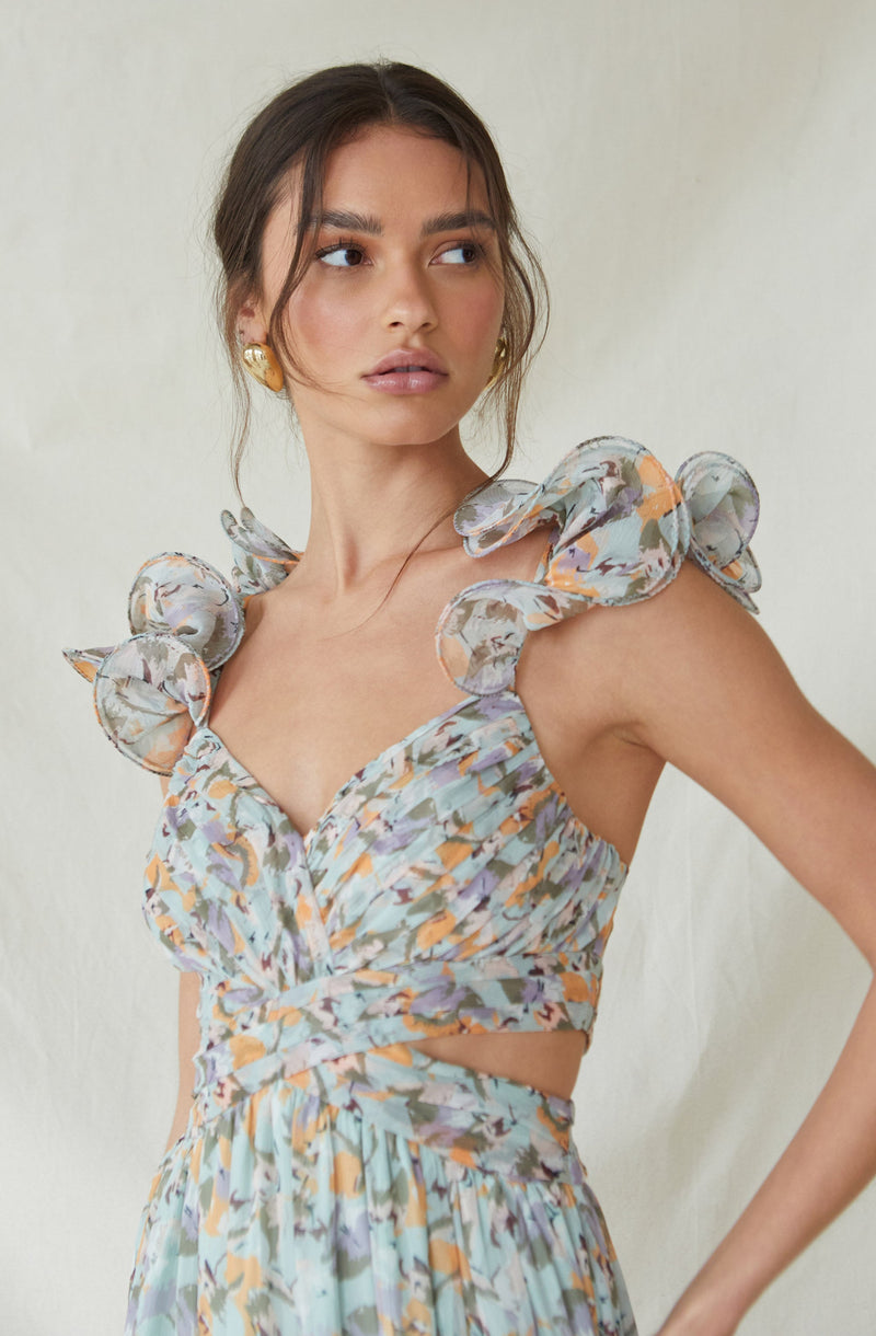 A young woman is wearing a sleeveless, floral-print dress with ruffled shoulder details. The dress features a fitted bodice with a v-neckline, a cut-out detail at the waist, and a flowing, pleated skirt. The fabric has a multicolored abstract floral pattern with shades of blue, purple, orange, and beige.