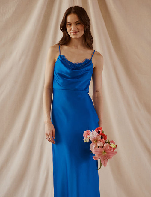 Woman in a blue satin slip dress with thin spaghetti straps and lace trim, holding pink flowers.