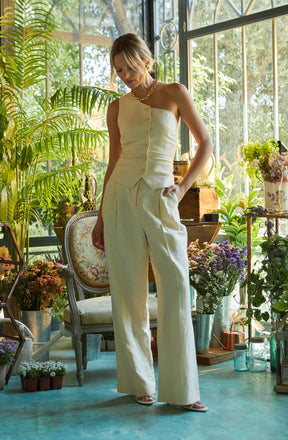 A woman is wearing a sleeveless, one-shoulder cream-colored top with button details down the front, paired with high-waisted, wide-leg cream-colored trousers. She accessorizes with a chunky gold chain necklace and light-colored open-toe shoes. The setting is a bright, plant-filled room with large windows, lush greenery, and various potted plants and flowers surrounding her.