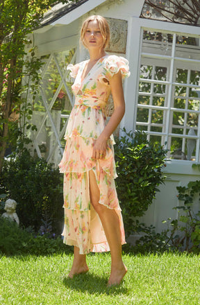 A woman is wearing a light, flowing dress with a floral pattern in pastel colors, featuring ruffled cap sleeves and a tiered skirt with a high slit.