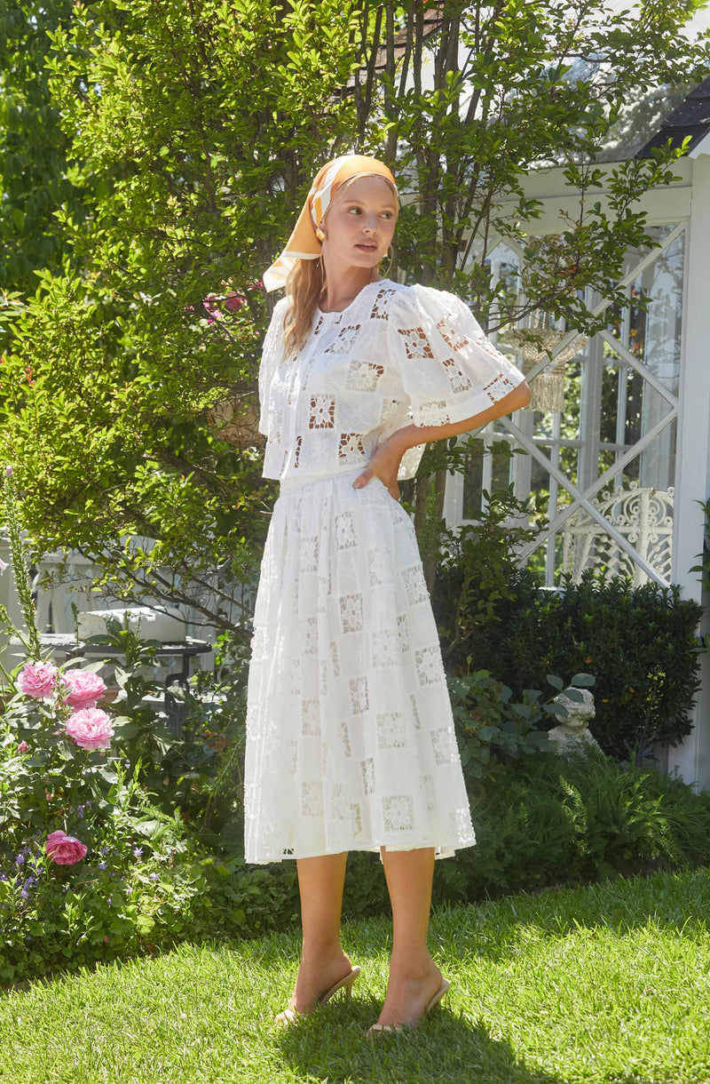 A woman is wearing a white dress made of a lightweight, eyelet fabric with square cut-out patterns. The dress has a fitted waist, a mid-calf length skirt, and a short-sleeved, loose-fitting top with a round neckline. She is also wearing a matching yellow and white headscarf, and nude-colored heeled sandals. The background features lush greenery, pink flowers, and a white garden structure.