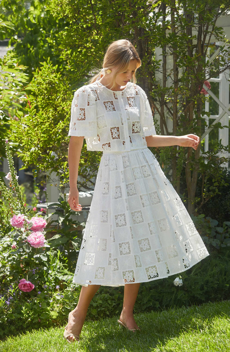 A woman is wearing a two-piece white dress with a square pattern made of eyelet lace. The top has short sleeves and a front button closure, while the skirt is long and flared. She is standing outdoors in a garden with blooming pink roses and lush green foliage.