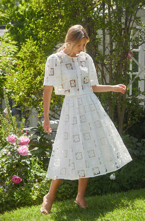 A woman is wearing a two-piece white dress with a square pattern made of eyelet lace. The top has short sleeves and a front button closure, while the skirt is long and flared. She is standing outdoors in a garden with blooming pink roses and lush green foliage.