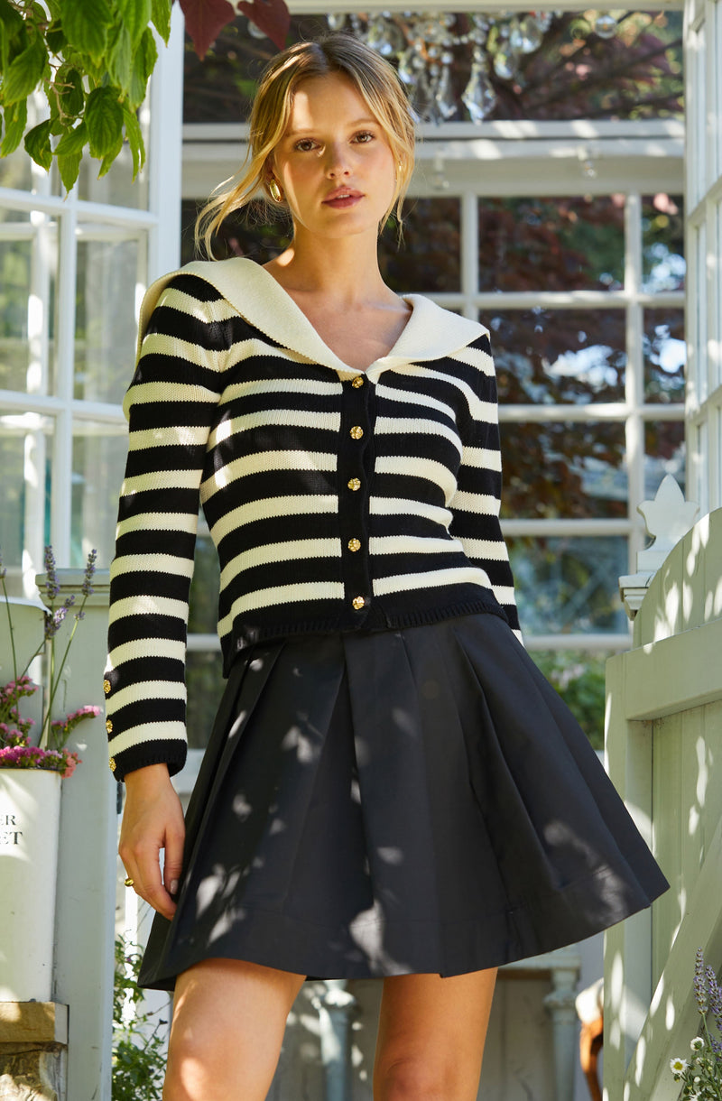 A woman is wearing a black and cream horizontal striped cardigan with gold buttons down the front and a scalloped collar. She is also dressed in a high-waisted black pleated skirt. The setting appears to be a sunlit greenhouse or conservatory with large windows and greenery around her.