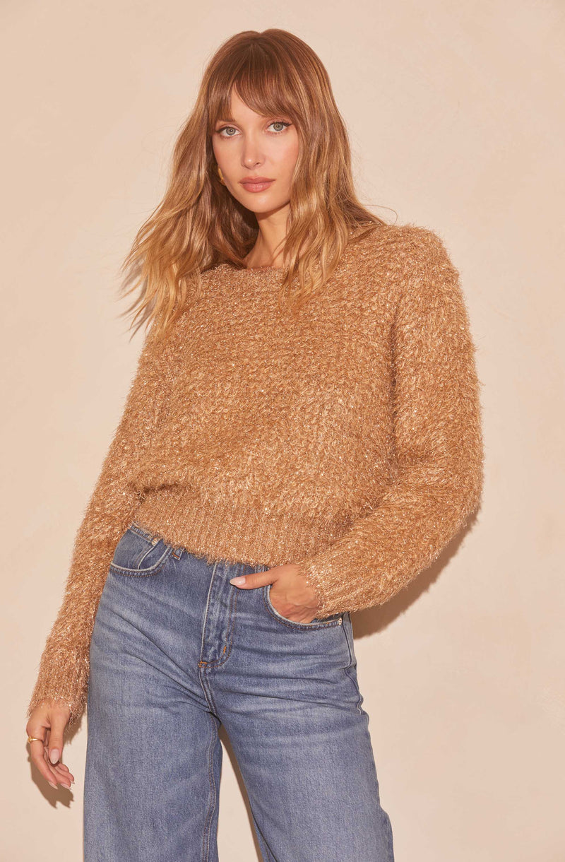 A woman is wearing a chunky, textured, light brown sweater with long sleeves and a round neckline. She has paired it with high-waisted, light blue jeans.