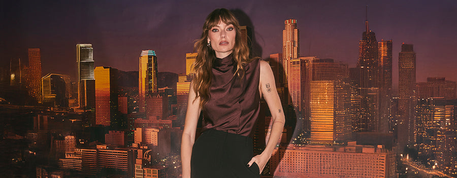 A woman stands in front of a city skyline at dusk, wearing a sleeveless, dark satin top with a high collar and a loose fit, paired with high-waisted black pants.