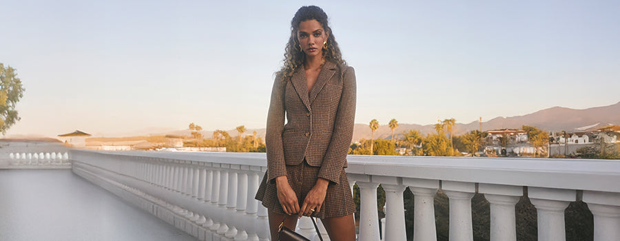 The woman is wearing a brown blazer over a brown dress with a subtle checkered pattern. She is holding a small handbag with a handle in front of her.