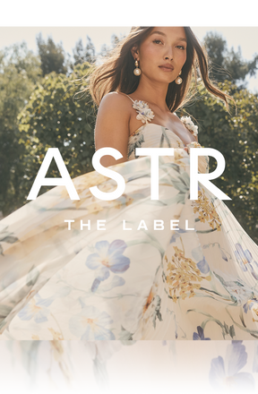 A woman is wearing a sleeveless, flowy dress with a floral print in soft pastel colors. The dress has a fitted bodice with decorative floral shoulder straps. The background features trees and a sunny sky.