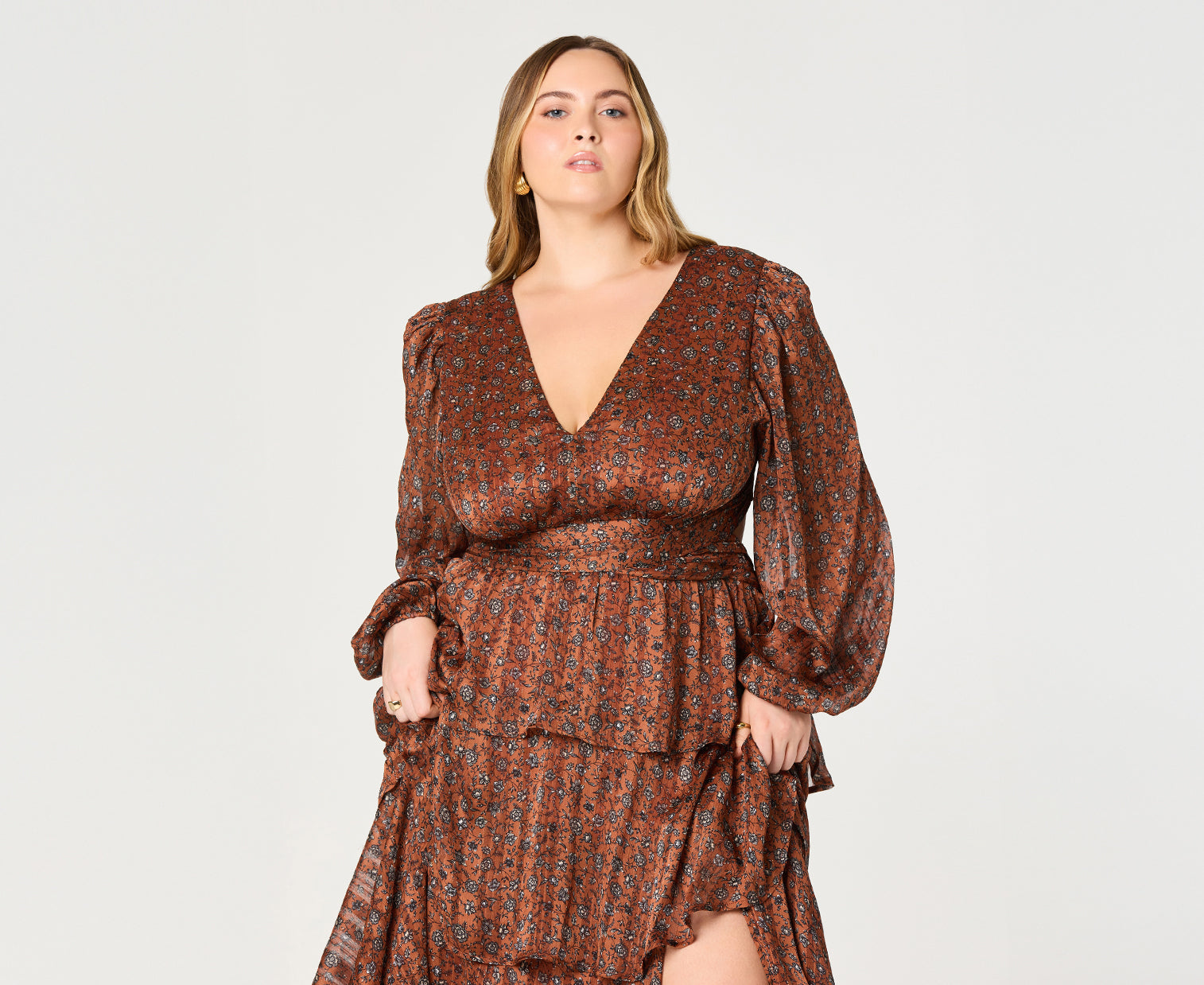 A woman is wearing a long, flowing dress in shades of brown with a small floral pattern. The dress features long, puffed sleeves with elastic cuffs, a deep V-neckline, and an elastic waistband. The skirt of the dress has a tiered, ruffled design with a slit on the side.