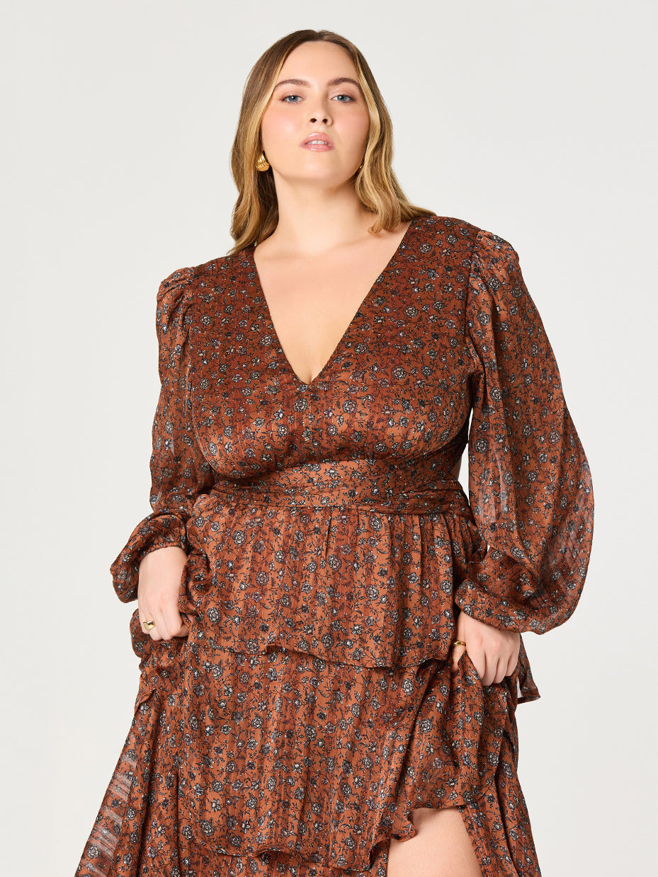 A woman with wavy blonde hair is wearing a long-sleeved, rust-colored dress with a small floral pattern in black and gray. The dress features a deep V-neckline, puffed shoulders, and gathered cuffs. The waistline is defined, and the skirt has layered ruffles, with one of the layers slightly lifted to reveal a slit.