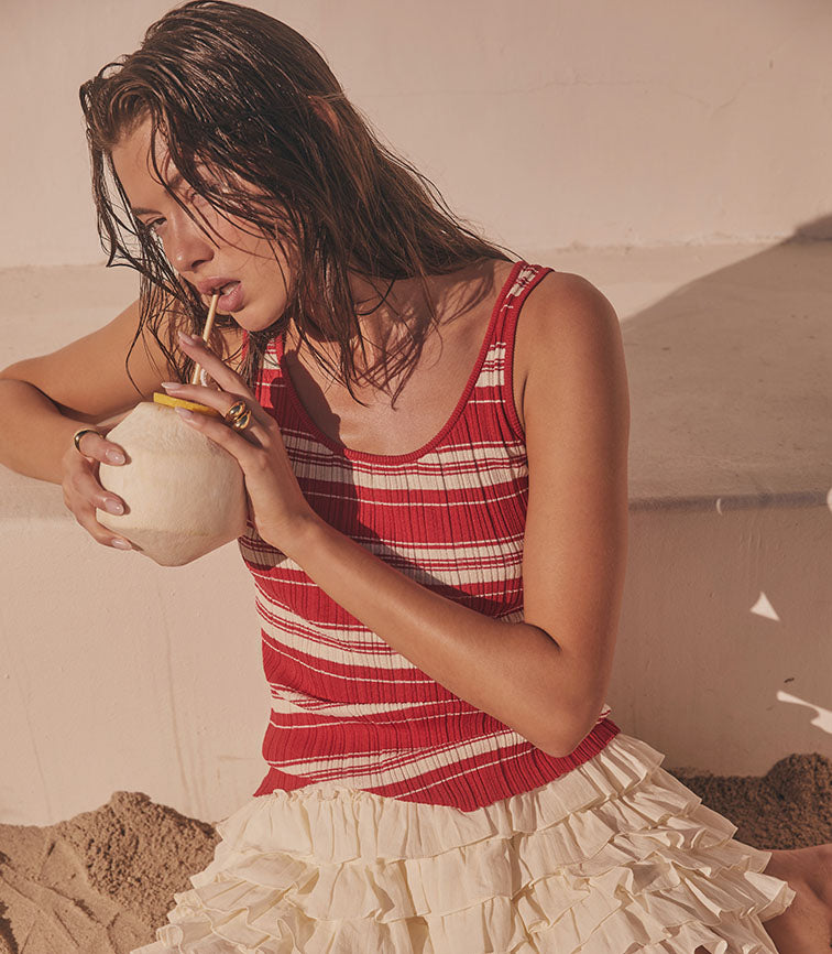 Woman in a red and white striped tank top and beige ruffled skirt, drinking from a coconut.