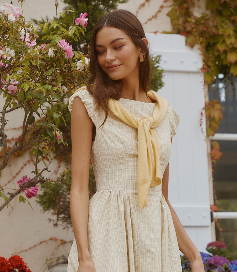 Woman wearing a light yellow dress with a matching scarf tied around her shoulders, outdoors among flowers.