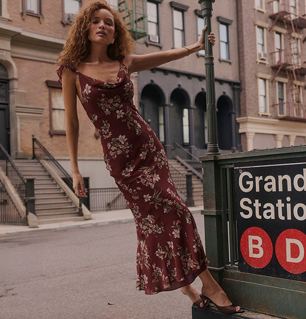 Woman in a long, floral burgundy dress standing next to a subway entrance.