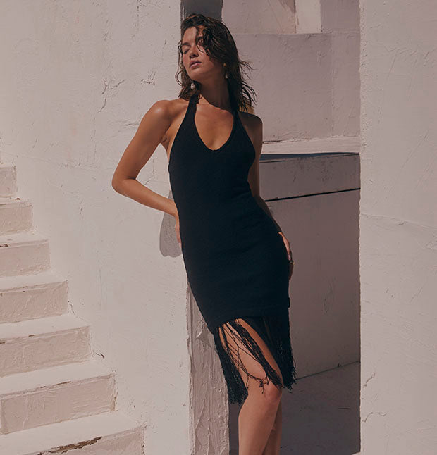 Woman in a sleeveless black dress with fringe detail at the hem, standing against a white wall.