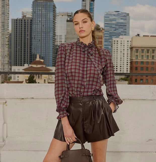 Young woman in a plaid long-sleeve blouse with ruffled collar and cuffs, and high-waisted black leather shorts, holding a handbag, on a rooftop.
